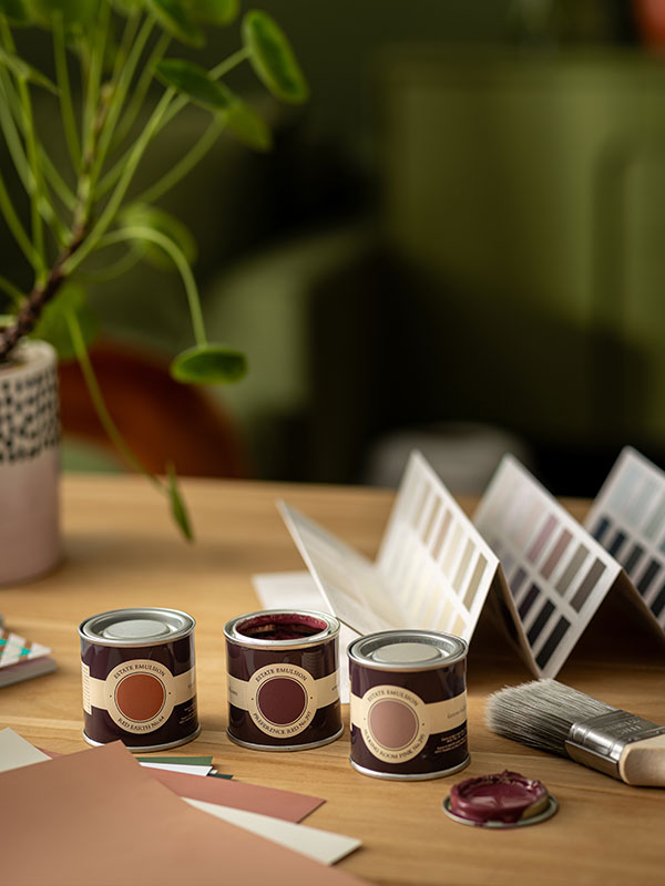 Three small paint tins in warm colours sit on a wooden desk with a paintbrush and colour cards.