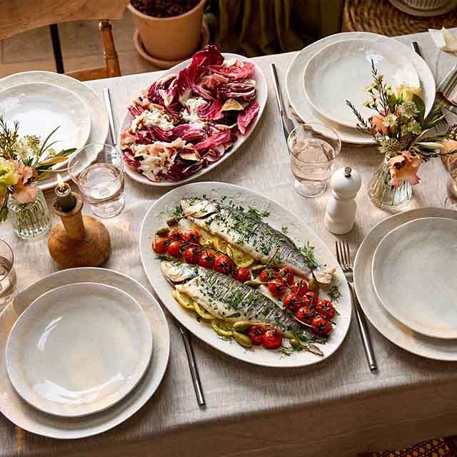 A table set for four with plates, glasses, two floral arrangements, and dishes of grilled fish with vegetables and radicchio salad.