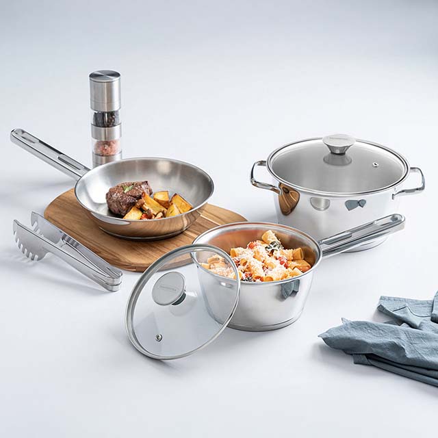 A set of stainless steel cookware with food, a spice grinder, and a blue napkin on a white background.