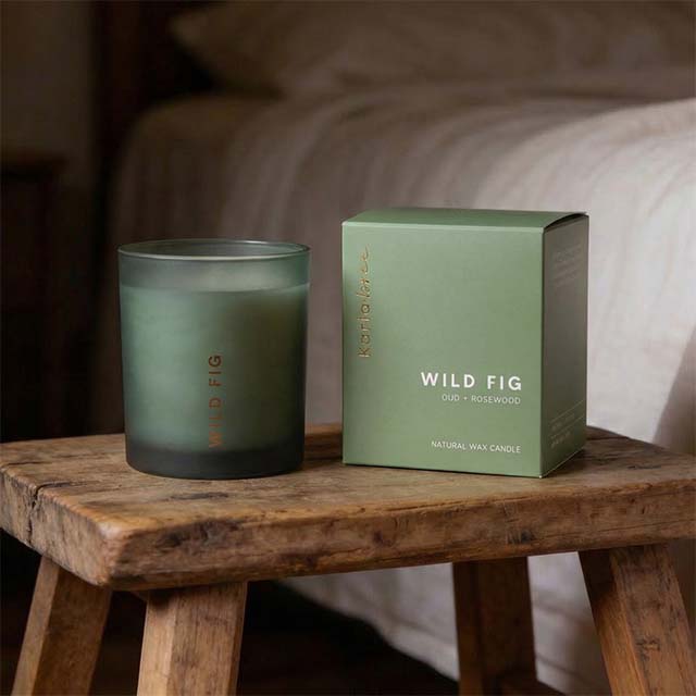 Green candle and box labeled 'Wild Fig' on a wooden table in a cozy room.