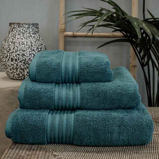 A stack of three teal towels on a wicker surface with a decorative vase and a plant in the background.