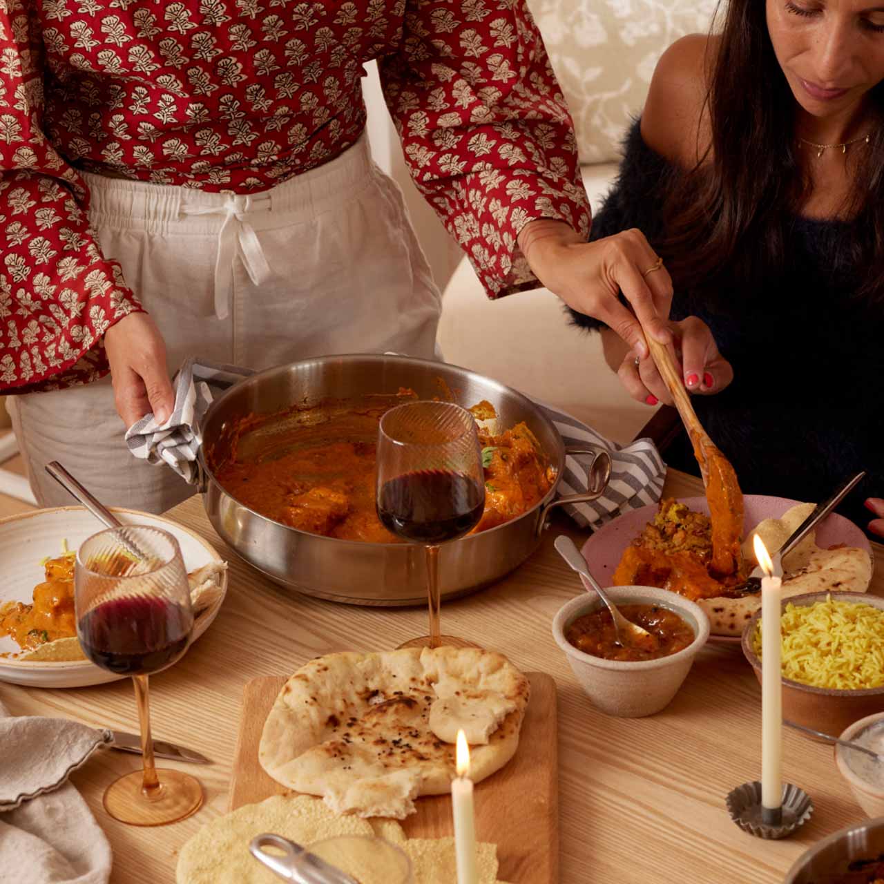 Two people enjoying an Indian meal with curry, naan, rice, and red wine on a wooden table.