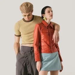 Hombre con gorra marrón de pata de gallo y camisa corta con pantalones grises, brazo alrededor de mujer con chaqueta de cuero roja, falda azul y botas beige.