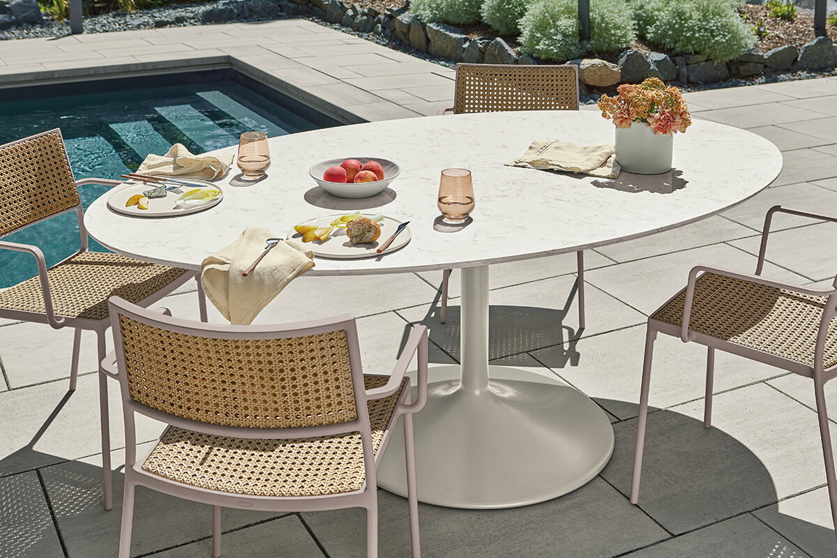 White Aria outdoor dining table with woven chairs beside a pool, set with plates, fruit and flowers.