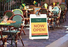 A sidewalk A-frame sign reading 'Now open' sits outside a cafe with outdoor seating and greenery in the background.