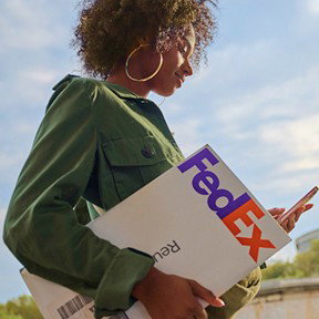 A smiling woman in a green jacket holds a FedEx box while looking down at her cellphone.