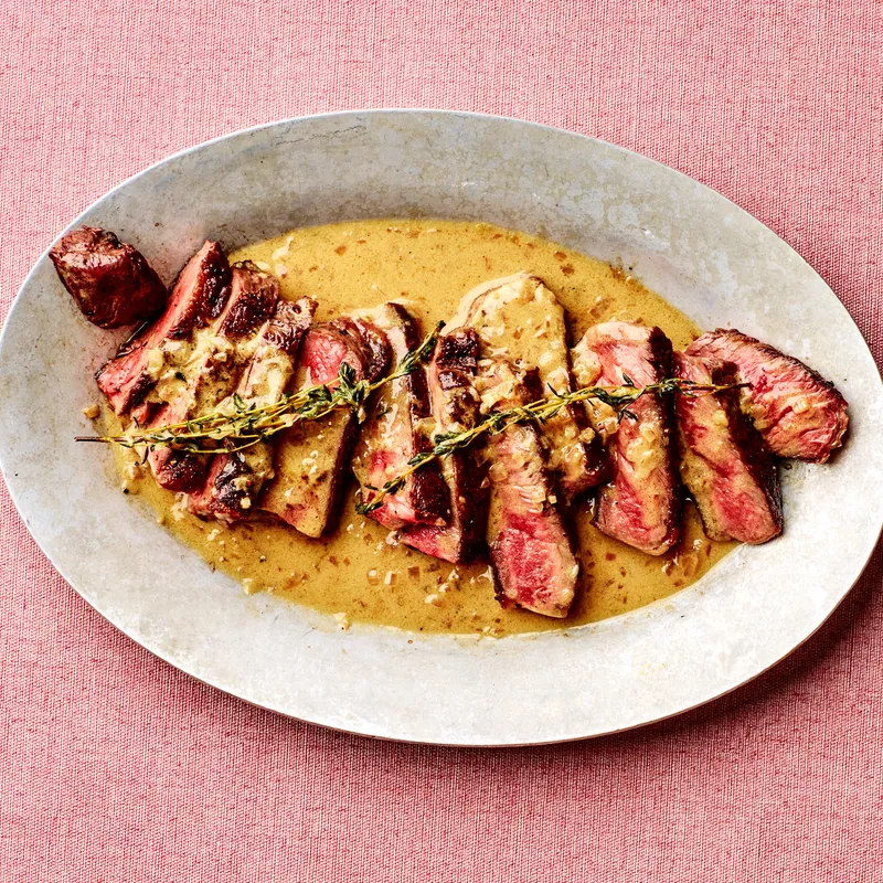 Sliced strip steak on a platter, dressed in pan sauce and garnished with thyme sprigs.