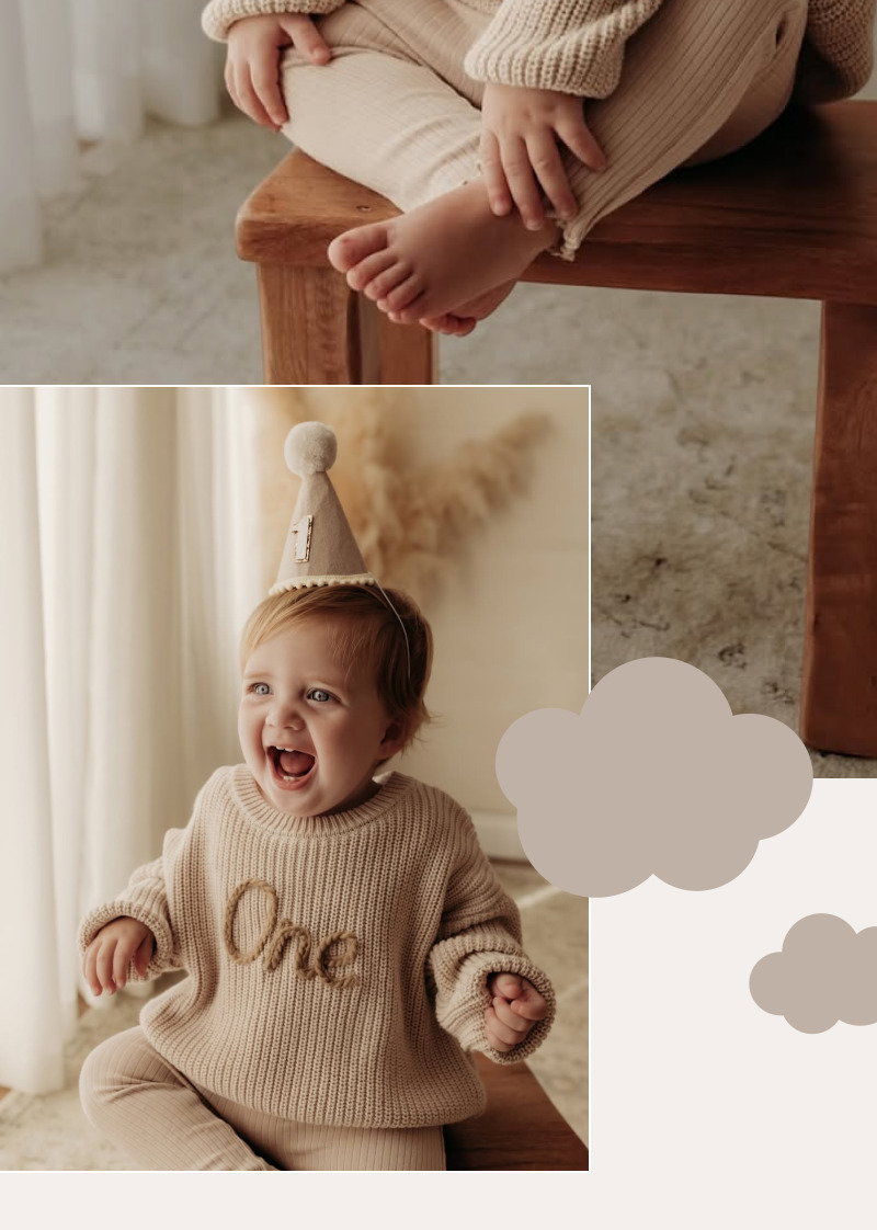 Smiling toddler wearing a beige knit sweater and light pants while sitting near a softly lit window indoors.