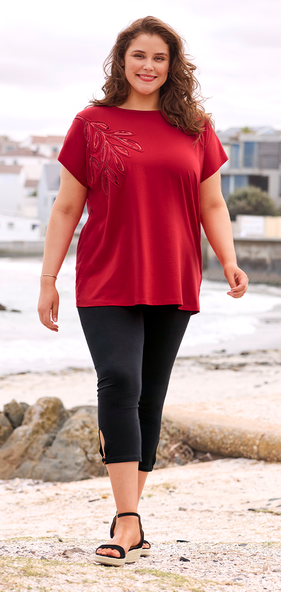 Frauenmodel trägt rotes T-Shirt mit U-Bootausschnitt und schwarze Leggins mit dekorativem Ring-Detail, Vorderansicht.