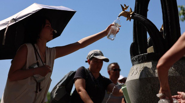 People wearing caps and holding umbrellas fill water bottles at a fountain in London on Monday against a bright blue sky.