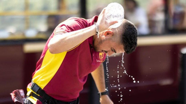 A man wearing a red and yellow uniform pours water from a bottle over his neck, which runs off his head and past his face as he looks down. The background behind him is blurred.