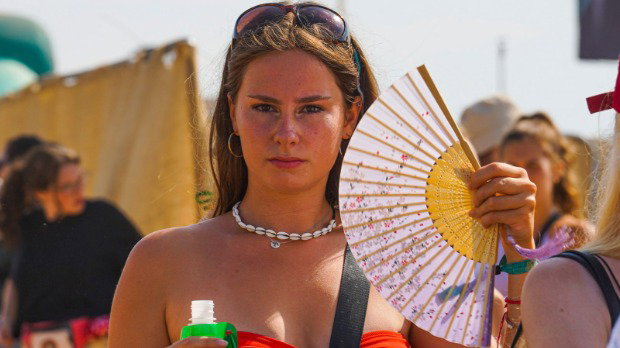 A woman holds a paper fan open and looks at the camera. She wears sunglasses on her head and wears a strapless top. The festival background is blurred.