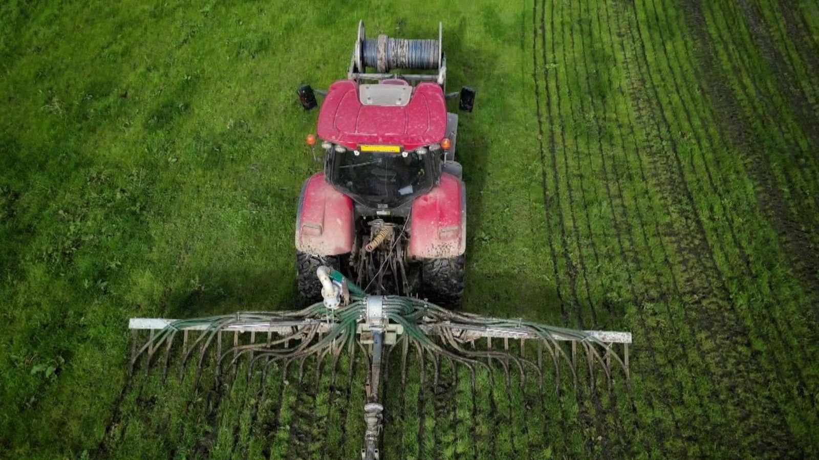Aerial view shows a red tractor with a large horizontal attachment spreading slurry in multiple rows at the back as it drives across a green field.