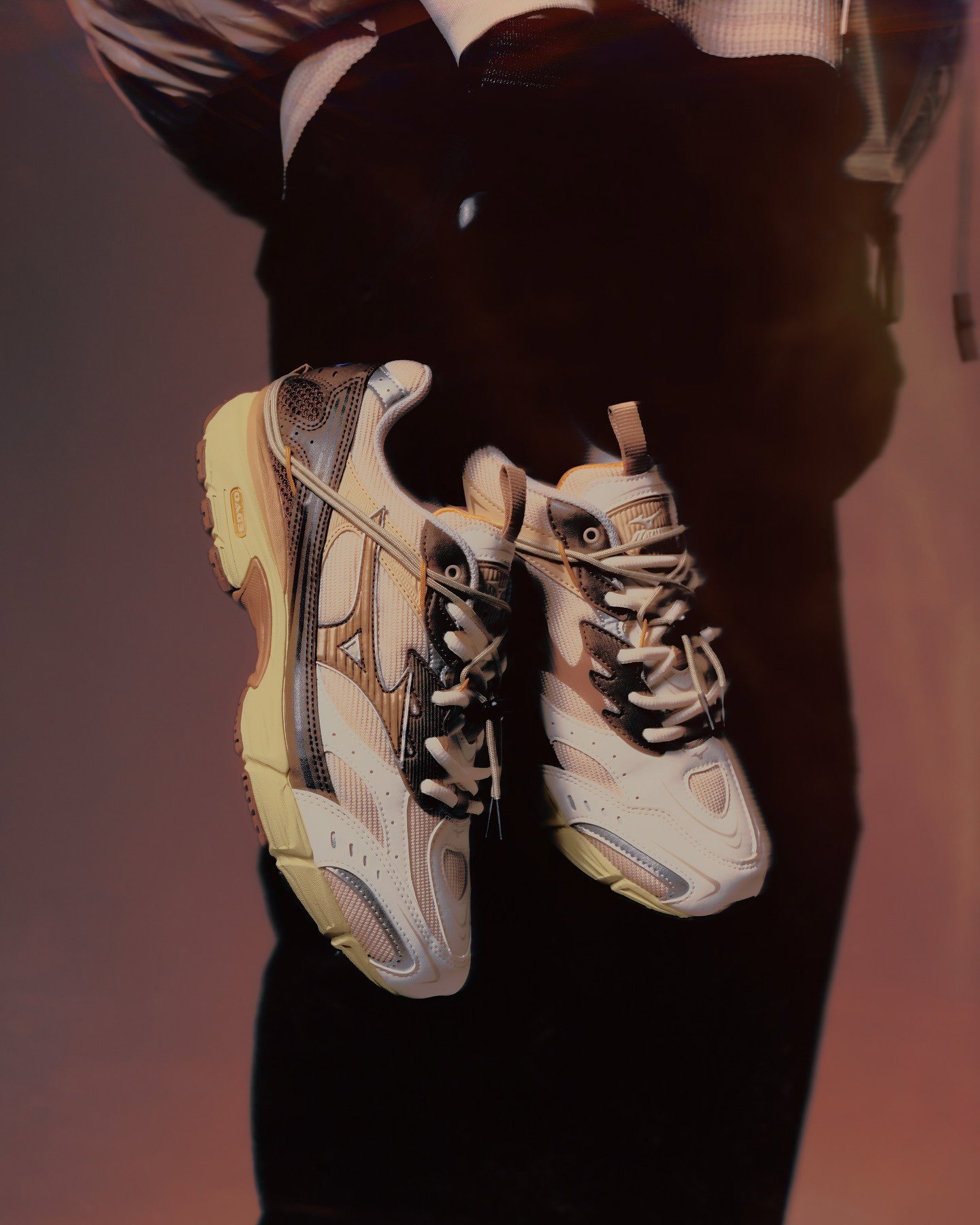 Pair of beige and brown chunky trainers hanging by their laces against a dark background, cropped person in frame