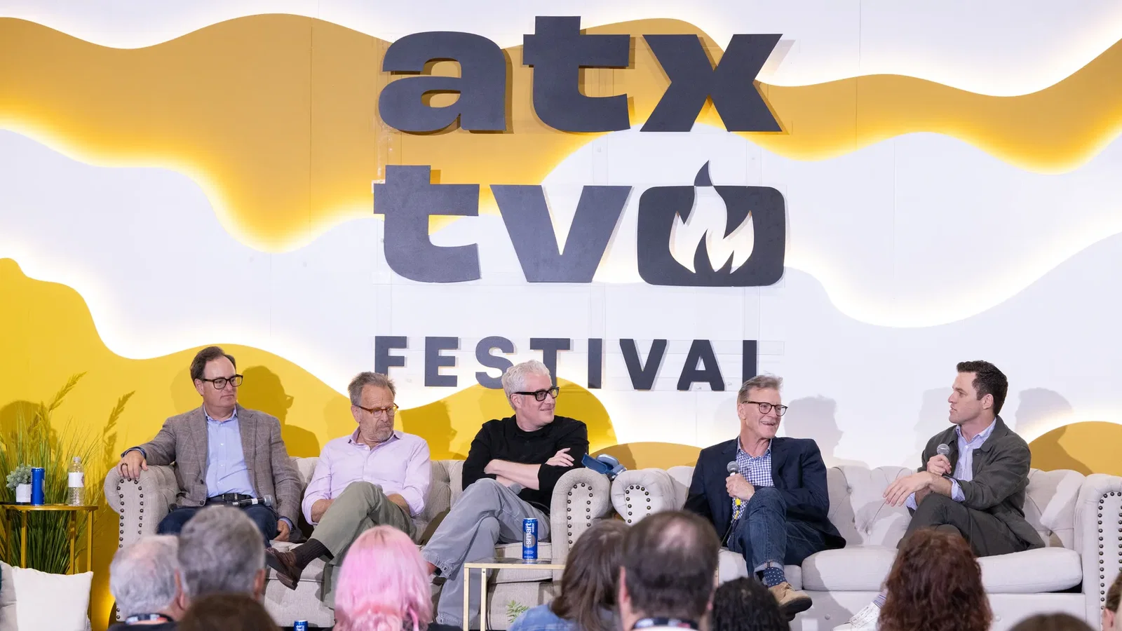 Panel discussion on a stage at the ATX TV Festival; a yellow wavy backdrop with large signage reading ATX TV FESTIVAL. Five men sit on a couch, microphones in hand, audience visible in the foreground.