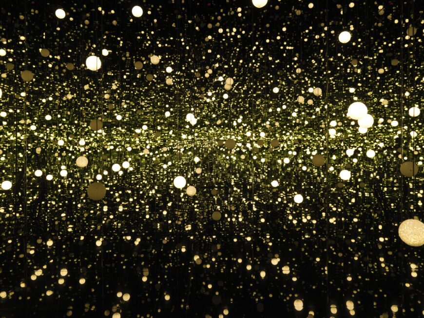 Against a dark background, hundreds of orbs of golden light appear to hover nearer or farther from our point of view. The orbs of light cluster toward a horizon line, making the distant center appear brighter.