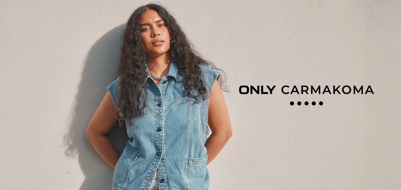 Vrouw met lang krullend haar, gekleed in een mouwloos denimvest en lichtblauwe broek, staande tegen een effen lichtgekleurde muur.