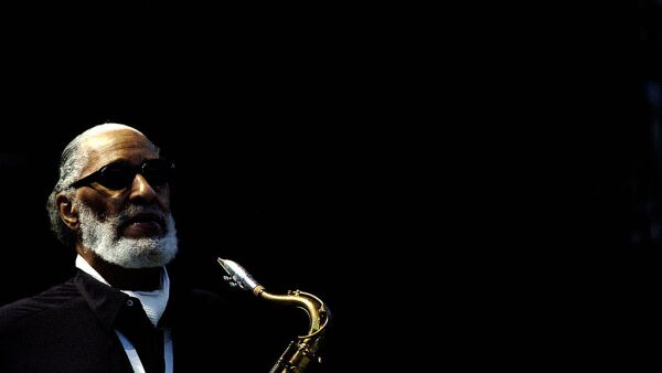 Sonny Rollins in 2006 holds a saxophone. He is wearing a black suit and black glasses. The background is black. His hair and beard are gray. 