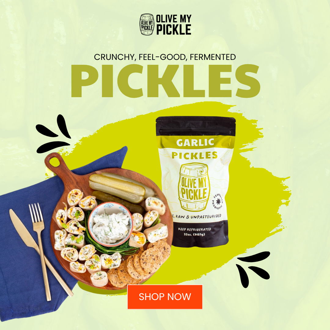 Pouch of Olive My Pickle Garlic Pickles next to a charcuterie board with sliced pickles, roll-ups, dip, and crackers on a blue napkin.