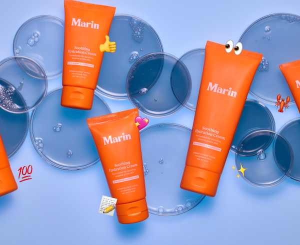 Alt text: Three bright orange Marin Soothing Hydration Cream tubes scattered across a light blue background with translucent petri dishes and bubbles. Playful emoji accents including thumbs up, sparkle hearts, eyes, lobster, sparkles, and “100” emoji surround the products.