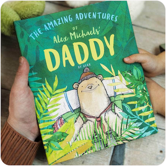 A young child holding up a personalised illustrated book titled "The Amazing Adventures of Dad"