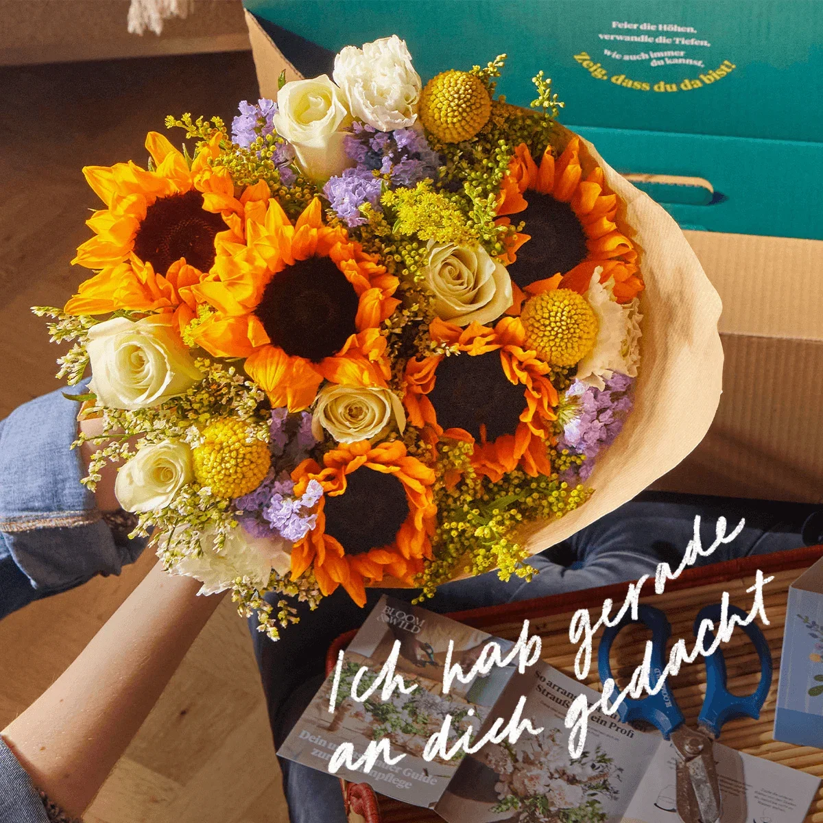 Ein sommerlicher Strauß aus leuchtenden Sonnenblumen, cremefarbenen Rosen, gelben Trommelstöckchen und fliederfarbenem Meerlavendel. Die Blüten wirken sonnig und texturreich. Weißer Text darüber sagt: „Ich hab gerade an dich gedacht“.