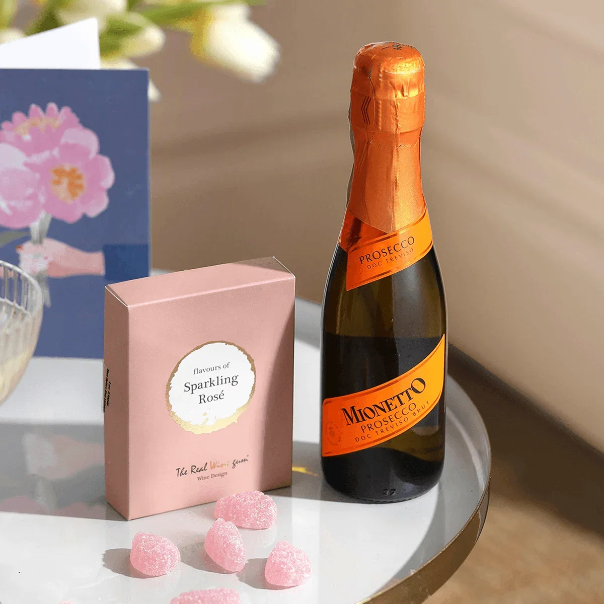 Ein festliches Geschenkset auf einem runden Tisch, bestehend aus einer kleinen Flasche Mionetto Prosecco und einer rosa Box Weingummis der Sorte „Sparkling Rosé“. Davor liegen zuckrige, rosa Fruchtgummis. Im Hintergrund lehnt eine Grußkarte mit Pfingstrosen.