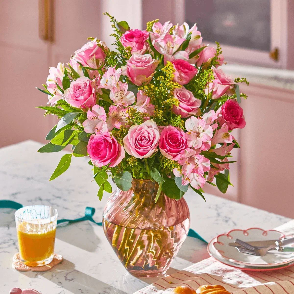 Eine geriffelte Glasvase ist mit einem romantischen Strauß aus rosa Rosen , zarten Inkalilien und gelben Goldruten  gefüllt. Eukalyptus  verleiht eine frische Note. Der Strauß steht auf einem hellen Tisch in einer freundlichen, hellrosa Küche.