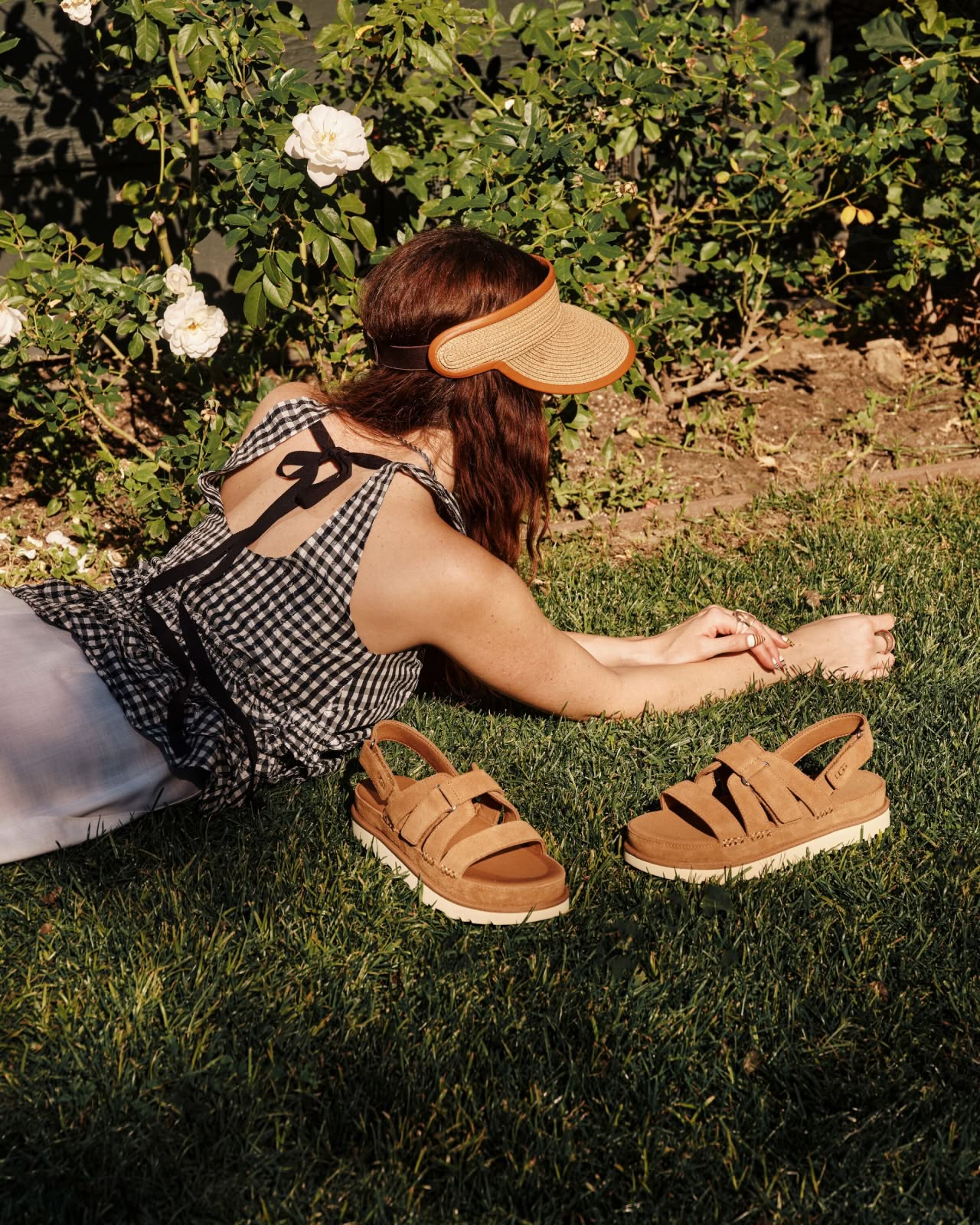A Person laying on grass, wearing polkadot top, straw visor and shorts with the goldenstar gleam sandal next to her.