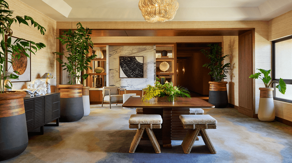Refined private lounge with warm wood detailing and greenery surrounding a central table beneath a statement chandelier.
