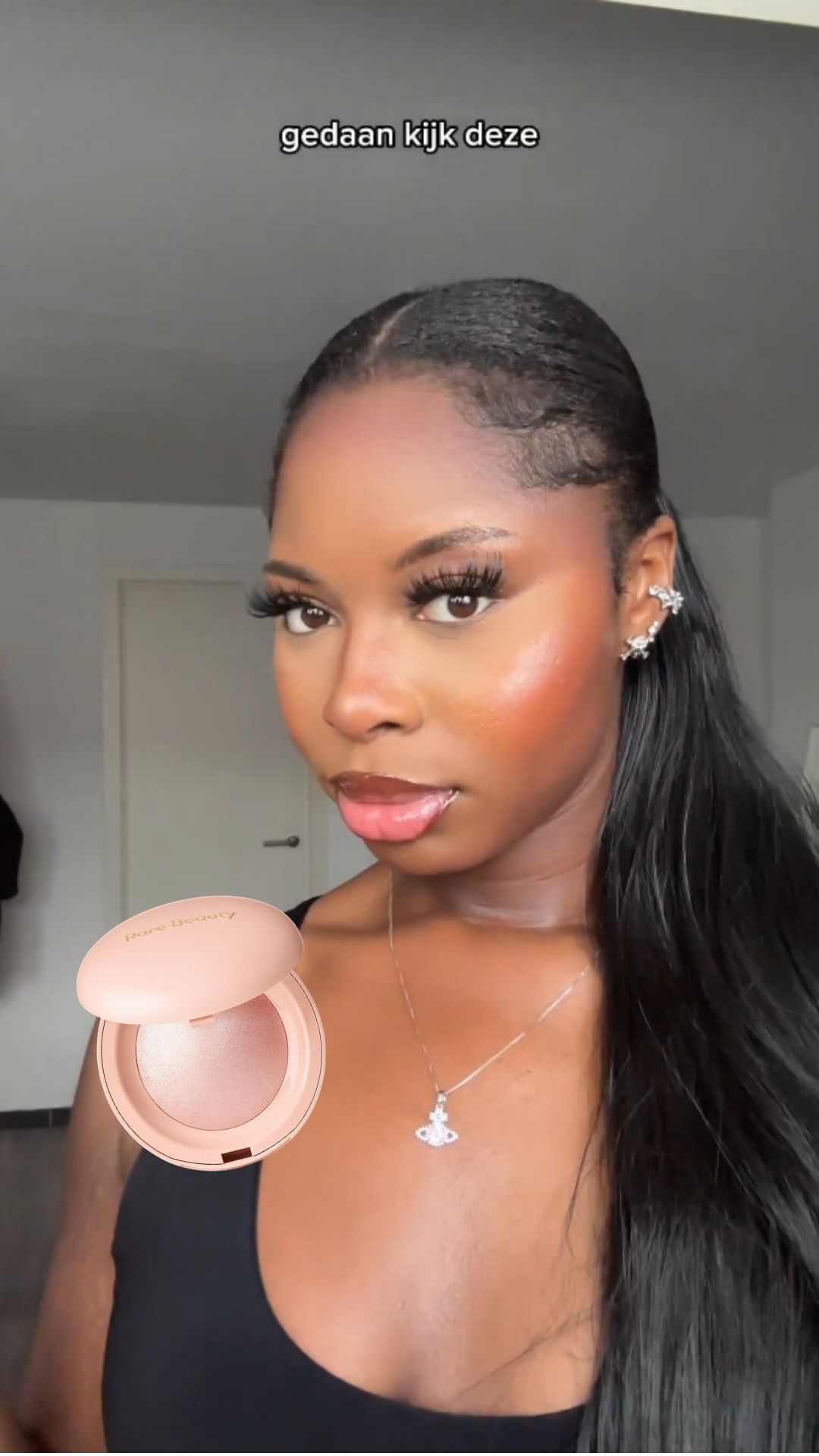 Een vrouw met lang haar en een zwarte top laat een Rare Beauty highlighter in een roze verpakking zien.