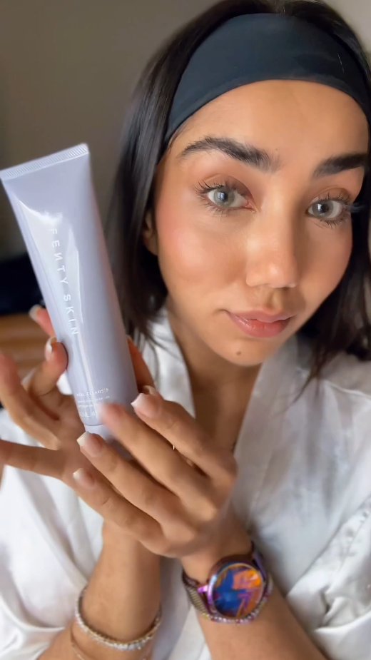 Een vrouw met een zwarte hoofdband houdt een lichtpaarse tube Fenty Skin gezichtsreiniger vast, klaar om je huid te laten stralen.