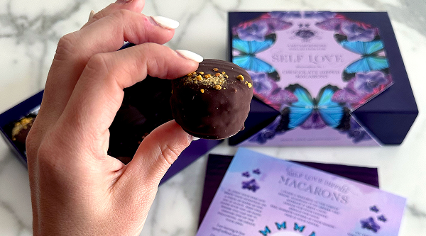 A hand holds a chocolate dipped macaron in front of the Vosges Self Love box