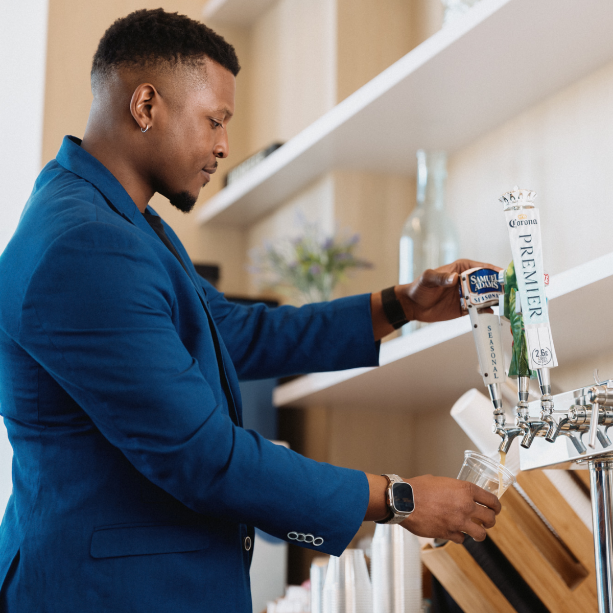 A bartender in a blue blazer pours draft beer from a tap into a clear plastic cup. He stands at a modern bar with metal beer taps and stacked cups visible in the foreground.