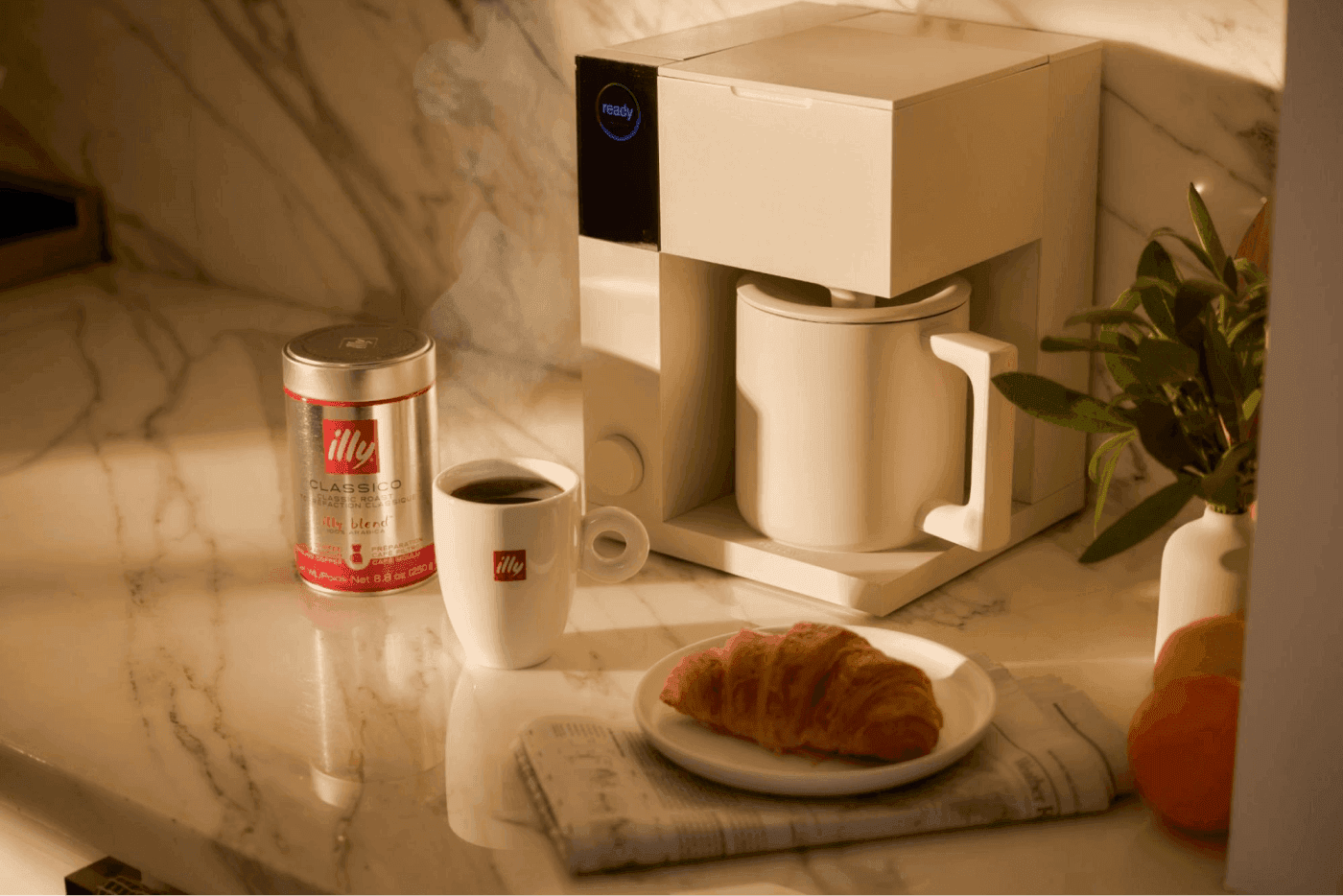Coffee maker with fresh coffee, croissant, and Illy coffee tin on marble counter