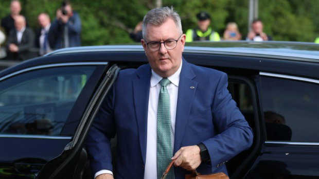 Sir Jeffrey Donaldson exits the back of a black car holding a brown leather satchel. He wears glasses, a dark blue blazer, green tie and white shirt. There is a small crowd behind him, including a police officer and someone taking a picture.