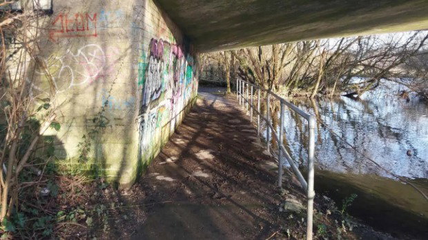 A footpath beside a river in Hampshire, leading under a road bridge. There is graffiti on the walls and white railings separating the pavement from the lake.