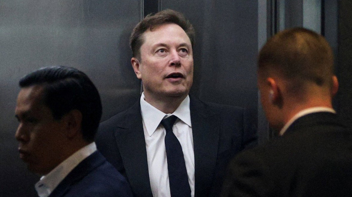 Elon Musk is stood looking into the distance as other people walk past him. He wears a black tie and black suit jacket.