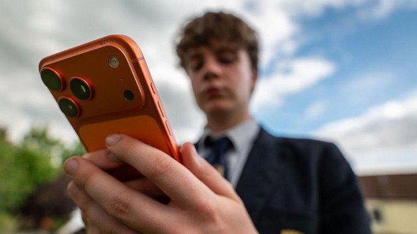 A boy wearing a school uniform holds an orange mobile phone in his hands. His face is blurred, and the phone is in the foreground. There is a blue cloudy sky behind him.