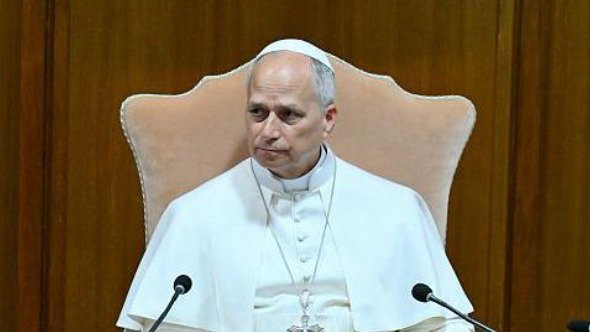 Pope Leo sits in a chair wearing white robes and a white hat, with two microphones pointed up at him from below. The background is a wooden-panelled wall.
