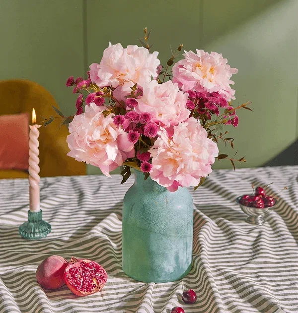A textured teal vase holds a lush bouquet of fluffy, pale pink peonies, fragrant eucalyptus sprigs, and deep magenta button chrysanthemums. Set on a striped tablecloth with a lit candle, a halved pomegranate, and fresh cherries.