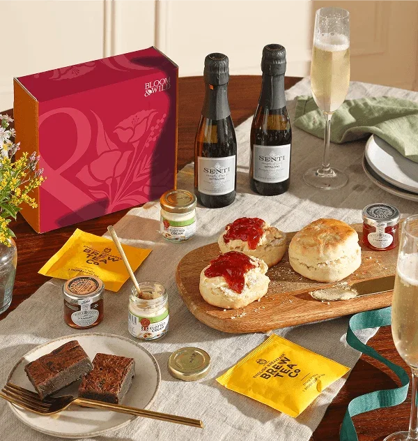 An indulgent Bloom & Wild afternoon tea gift hamper. Features fresh, crumbly scones topped with thick clotted cream and glossy strawberry jam, rich chocolate brownies, Brew Tea Co bags, mini Senti Prosecco bottles, and a fluted glass of fizz.