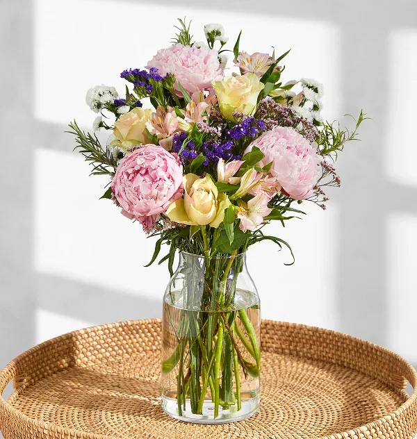 A vibrant Bloom & Wild bouquet with ruffled bright pink peonies, peach roses, lilac alstroemeria, and fragrant rosemary stems. Displayed in a striped ceramic jug on a coffee table, evoking a sunny, relaxed afternoon mood.