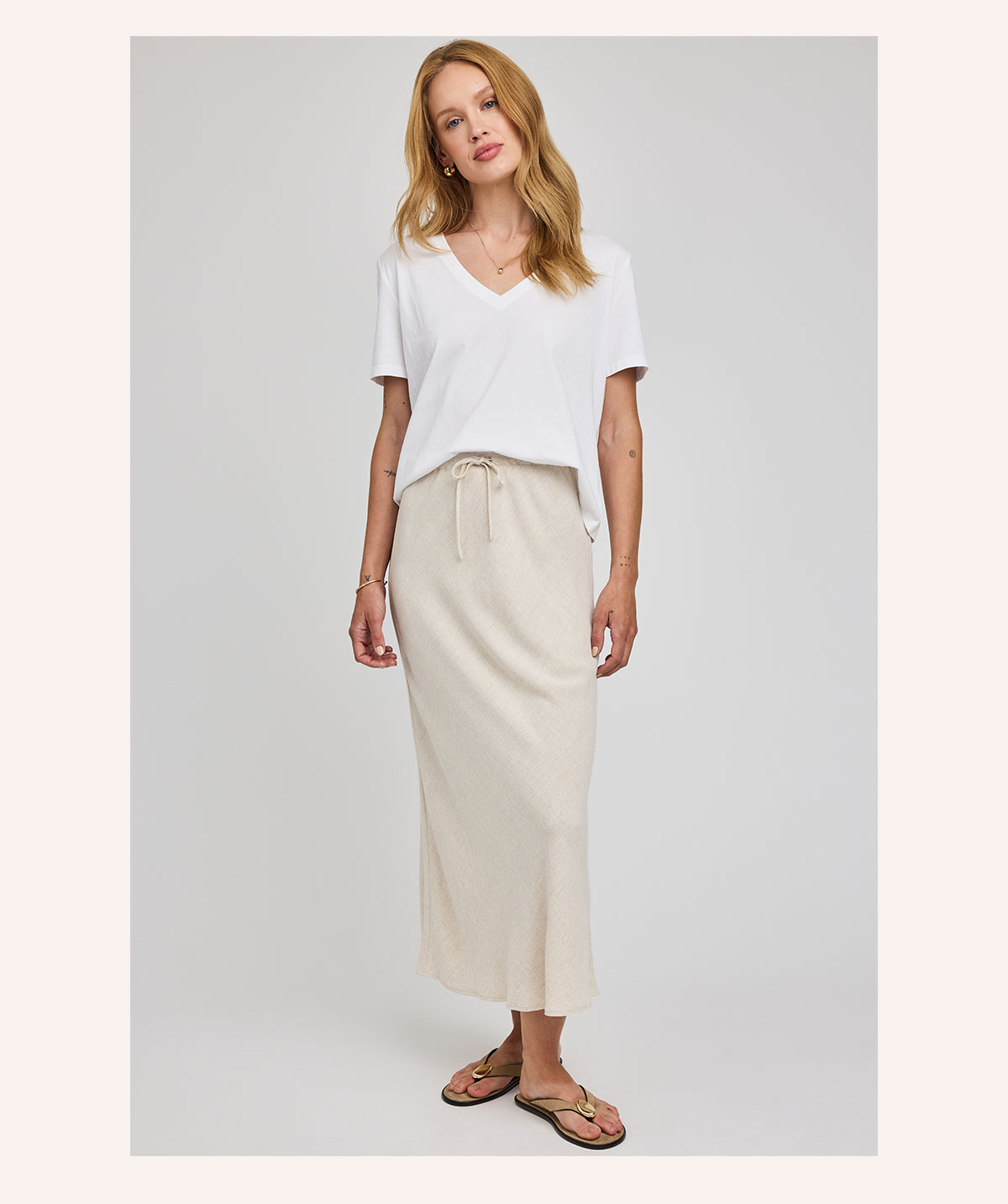 Model wearing a white v-neck t-shirt with a cream Cassia Skirt and sandals against a neutral studio background.