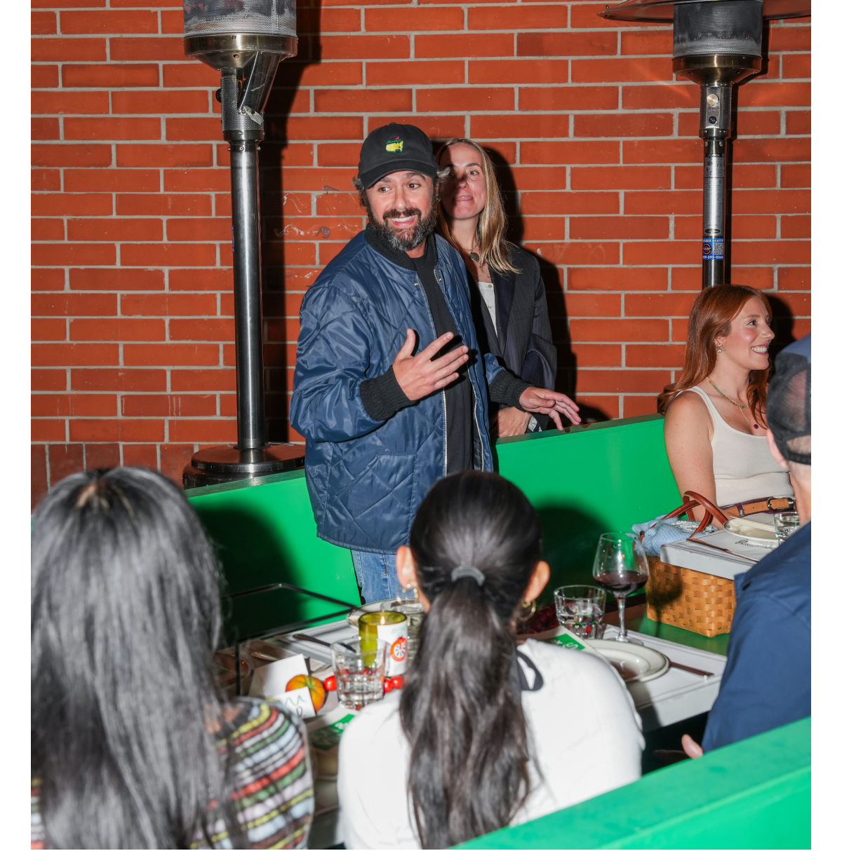 A guest addressing the group in front of a brick wall and outdoor heaters while attendees listen during the intimate Italian-inspired dinner gathering.