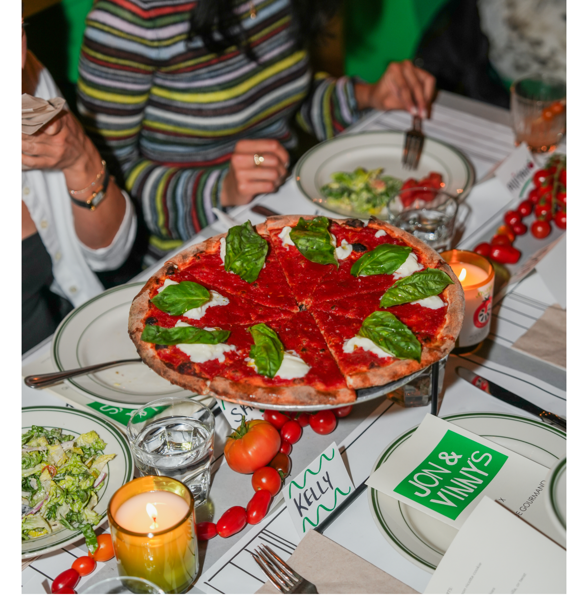 A restaurant table setting with a tomato and basil pizza, candles, cherry tomatoes, salads, and Jon & Vinny’s branded menus, capturing the atmosphere of a candlelit Italian-inspired dinner experience.