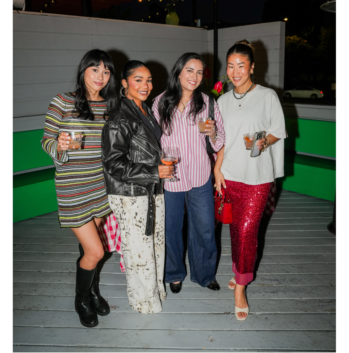 Four women posing together at an outdoor evening event holding drinks.