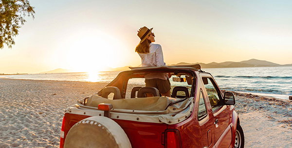 Frau steht im Cabrio am Strand und genießt den Sonnenuntergang
