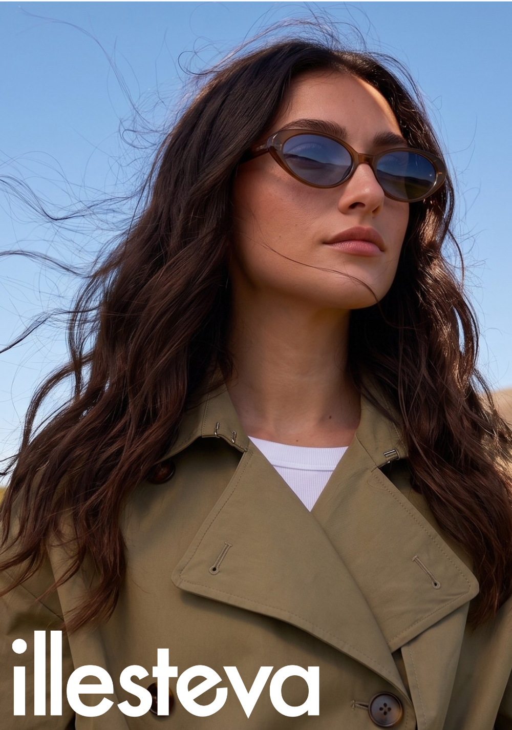 A campaign portrait of a woman looking off-camera against a clear blue sky. She has long, wavy dark hair blowing in the wind and is wearing a classic olive green trench coat over a white top. She is wearing the Sheila sunglasses, featuring a translucent brown, 90s-inspired oval silhouette. The white illesteva logo is positioned in the lower-left corner.