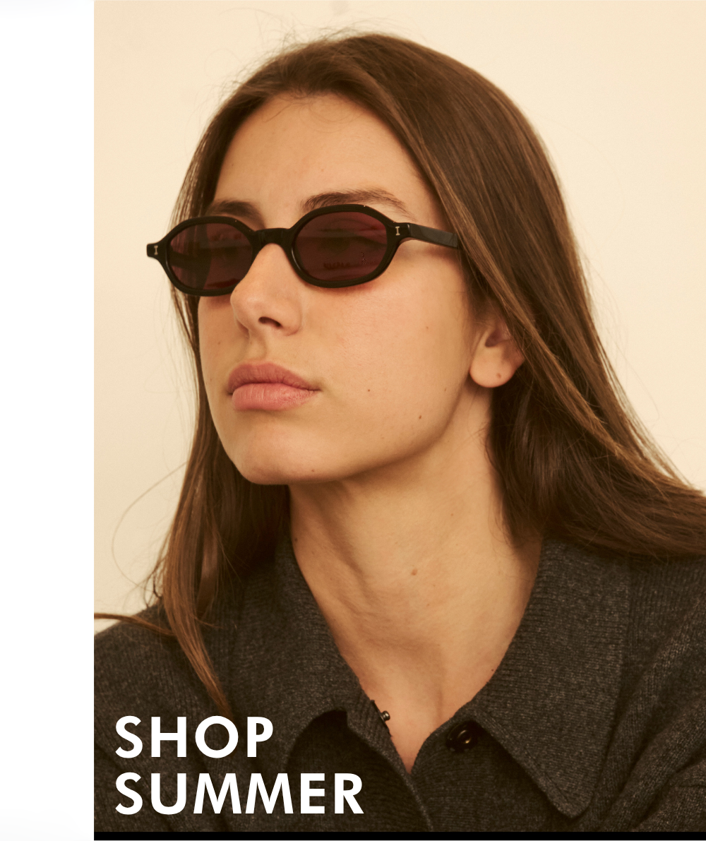 A lifestyle portrait of a woman looking to the side against a neutral background. She is wearing a dark grey textured button-up shirt and small, narrow black oval sunglasses with dark reddish-pink tinted lenses. Bold white text in the lower-left corner reads 
